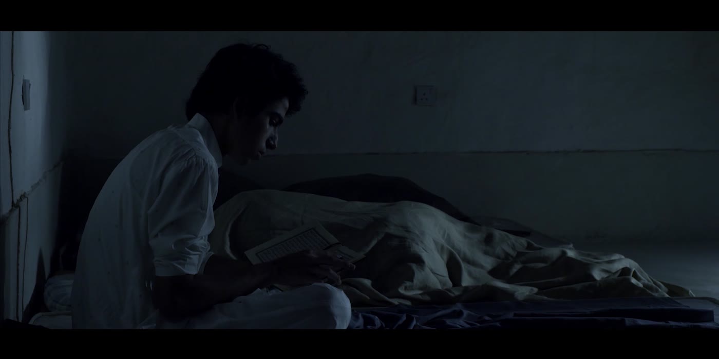 A young man leans over a sleeping figure in a dark room.