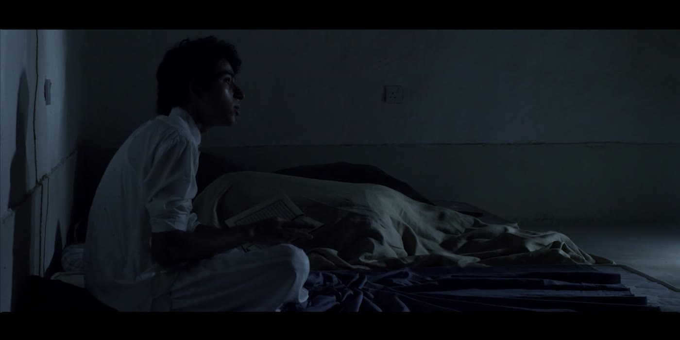 The young man sits upright in profile, waiting in low light.