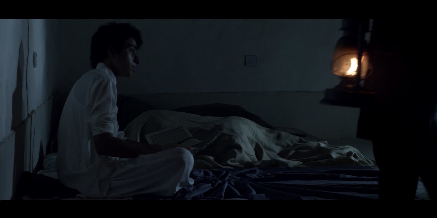 A warm lantern glows beside the young man in the darkened room.